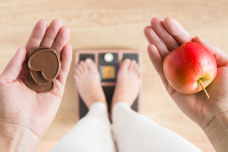 チョコレートとリンゴを持って体重計に乗る女性